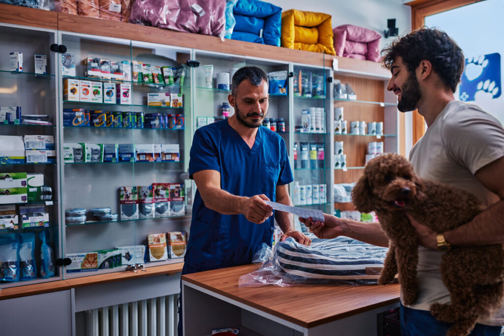 Veterinarian in blue uniform handing over a prescription and pet bed to a smiling customer holding his poodle in a vet clinic